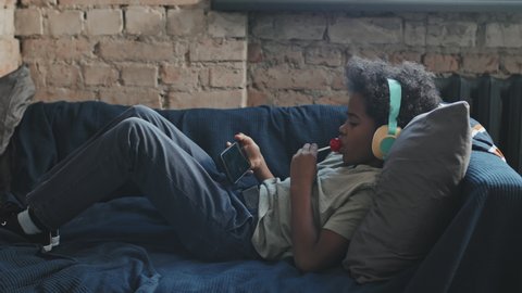 Full side view of ten-year-old Black boy wearing over-ear headphones, lying on couch in his room at daytime, having lollipop and using smartphone