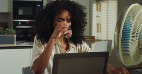 Cinematic shot of young brazilian business woman drinks glass of fresh water with switched on fan because of heat, doing smart working with laptop from home during covid-19 pandemic lockdown.