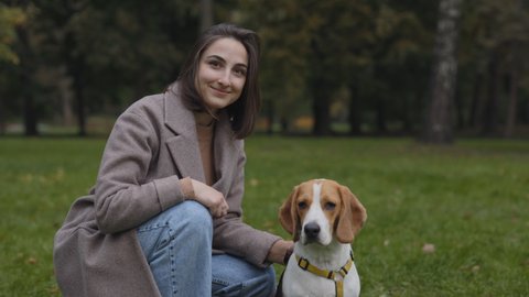 Charming dark haired woman looking at camera while sitting near her adorable beagle dog at city park. Owner and pet posing together outdoors.
