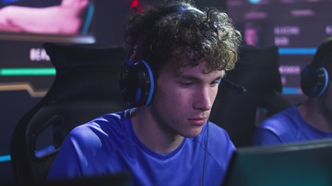 Concentrated esportsman gamer player with curly hair and headphones leaning back on chair in disappointment after failure battle in video game match during professional tournament