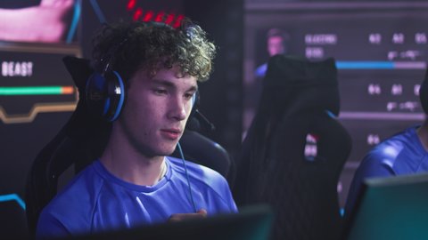 Serious esportsman gamer player with headset and curly hair looking at computer screen and sipping fresh water from bottle during break in gaming championship