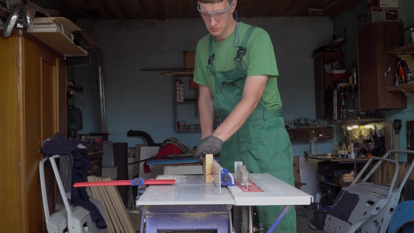 Carpenter in protective gloves and glases handles a wooden board works on planing machine in workshop.