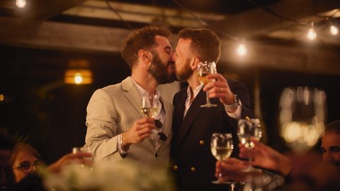 Big Dinner Party with a Crowd of Multiethnic Diverse Friends Celebrating at a Restaurant. Handsome Happy Gay Hosts Propose a Toast and Raise Wine Glasses while Sitting at a Table in the Evening.