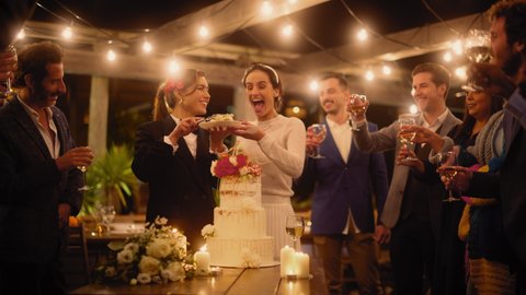 Beautiful Happy Lesbian Couple Celebrate Wedding at an Evening Reception Party with Diverse Multiethnic Friends. Queer Married Couple Standing at a Dinner Table, Kiss and Cut Wedding Cake.