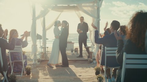 Beautiful Female Queer Couple Exchange Rings and Kiss at Outdoors Wedding Ceremony Near Ocean. Two Lesbian Women in Love Share Their Big Day with Diverse Multiethnic Friends. LGBTQ Relationship Goals.