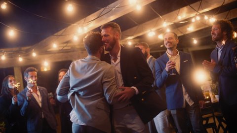 Handsome Happy Gay Couple Celebrate Wedding at an Evening Reception Party with Diverse Multiethnic Friends. Queer Newlyweds Dancing and Hugging at a Restaurant Venue. LGBTQ Relationship Goals.