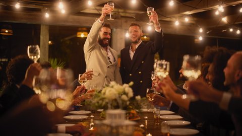 Big Dinner Party with a Crowd of Multiethnic Diverse Friends Celebrating at a Restaurant. Handsome Happy Queer Hosts Propose a Toast and Raise Wine Glasses while Sitting at a Table in the Evening.