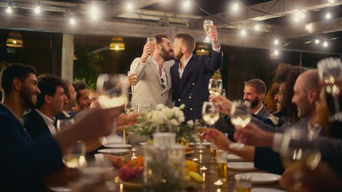 Big Dinner Party with a Crowd of Multiethnic Diverse Friends Celebrating at a Restaurant. Handsome Happy Hosts Propose a Toast and Raise Wine Glasses while Sitting at a Table in the Evening.