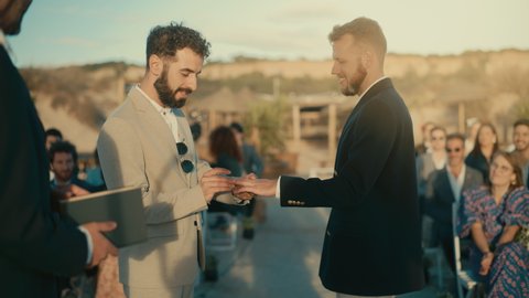 Handsome Gay Couple Exchange Rings and Kiss at Outdoors Wedding Ceremony Venue Near the Ocean. Two Happy Men in Love Share Their Big Day with Diverse Multiethnic Friends. LGBTQ Relationship Goals.