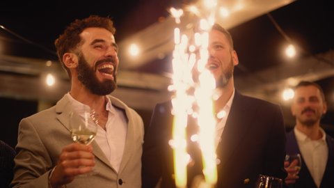 Handsome Happy Gay Couple Celebrate Wedding at an Evening Reception Party with Diverse Multiethnic Friends. Queer Newlyweds Kiss to Happy Marriage, Standing at a Dinner Table with Cake with Sparklers.