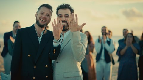 Portrait of a Happy Just Married Handsome Gay Couple Kissing, Showing Off Their Gold Wedding Rings. Two Attractive Queer Men in Suits Smile and Pose for Camera with Friends. LGBTQ Family Goals.