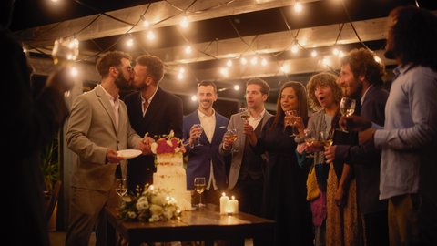Handsome Happy Gay Couple Celebrate Wedding at an Evening Reception Party with Diverse Multiethnic Friends. Queer Married Couple Standing at a Dinner Table, Kiss and Cut Wedding Cake.