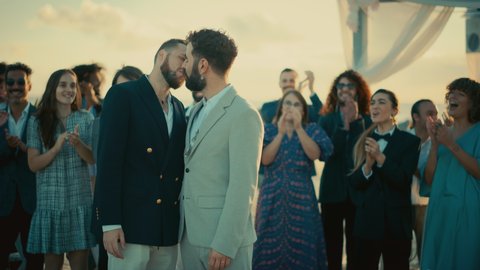 Portrait of a Happy Just Married Handsome Gay Couple Kissing. Two Attractive Queer Men in Suits Smile and Pose for Camera with Diverse Friends. LGBTQ Relationship and Family Goals.