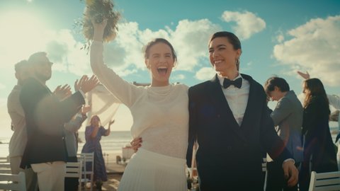 Beautiful Female Queer Couple Walking Up the Aisle at Outdoors Wedding Ceremony Near Sea. Two Lesbian Women in Love Share Happiness with Diverse Multiethnic Friends. Authentic LGBTQ Relationship Goals
