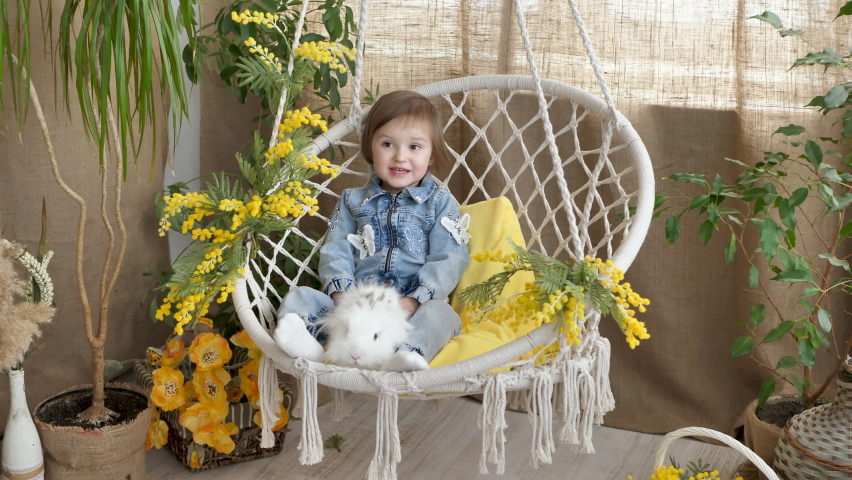 Little girl is riding in a wicker swing and stroking a white live rabbit on a festive Easter day.