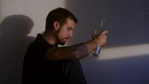 Upset young guy drinker alcoholic sitting at home with bottle drinking vodka alone