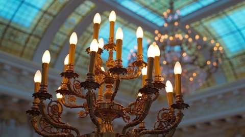 large gilded bronze candlestick with electric candles. luxury interior element. warm lights lamps against yellow blue royal style stained glass window hall and expensive crystal chandelier. close up