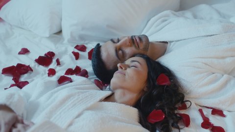 Beautiful Ethnic Couple Laying in Bed Full of Rose Petals, Sleeping As They Celebrate Valentines Day Together By Spending the Night in a Fancy Decorated Hotel.  Lovers Day Concept, Engagement.