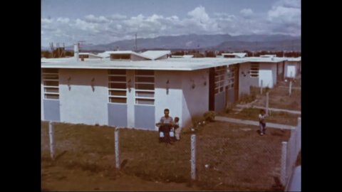 CIRCA 1960s - A boy helps to water his family's lawn while his father reads the paper in Guatemala.