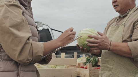 Mature farm worker demonstrating quality of vegetables to female customer standing in front of him making notes in papers