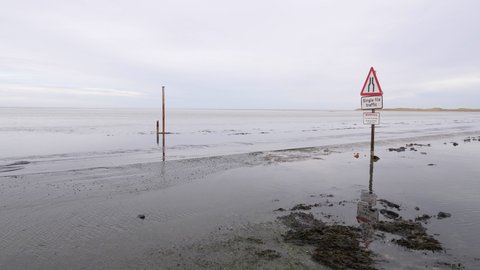 Lindisfarne , Northumberland , United Kingdom (UK) - 12 13 2021: Car driving along low flooded road past warning sign. Reflective water