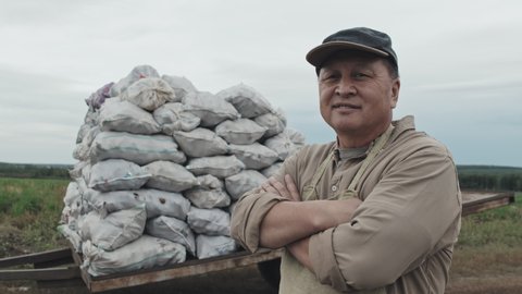 Horizontal medium portrait shot footage of confident mature Asian farmer standing with arms crossed against sacks of potatoes