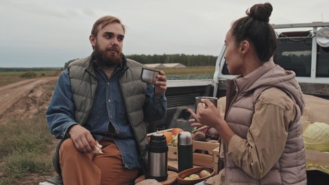 Young adult man and woman sitting relaxed on pickup truck cargo having lunch break during workday on farm field