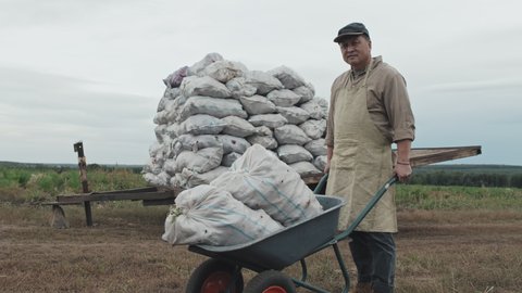 Medium long shot of mature Asian farm worker standing with cart with sacks of potatoes looking at camera