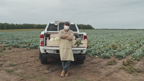 Zoom in shot footage of unrecognizable mature farm worker wearing mask standing with arms crossed leaning against pickup truck