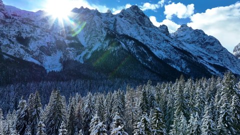 Beautiful drone view of Dolomites Mountains with ice and snow in winter, surrounded by forest with pines. Wide shot, daylight with blue sky and snow