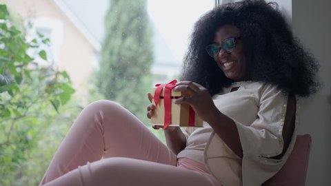 Surprised excited African American woman opening gift box sitting on windowsill smiling. Portrait of happy young beautiful millennial lady enjoying surprise indoors at home. Slow motion