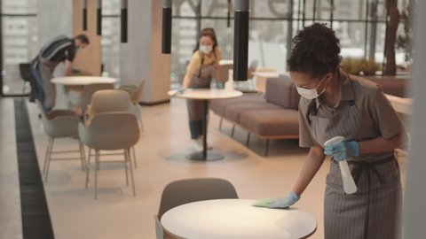 Steadicam of young Biracial woman wearing face mask and gloves, wiping table with disinfecting liquid in modern restaurant, her colleagues working on background