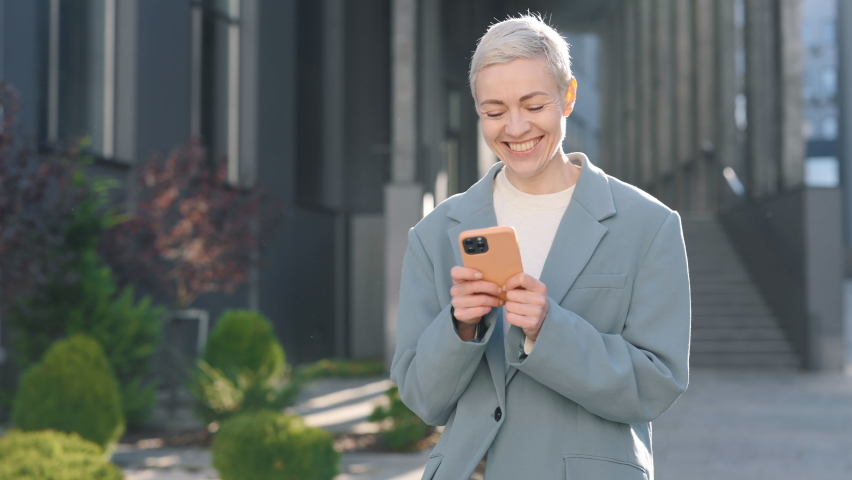 Smiling caucasian lady in formal wear texting on smartphone while walking outdoors near modern office center. Lifetyles, people and gadgets concept.