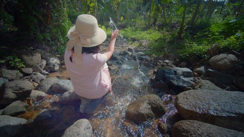 A female tourist sits on a rock in the middle of a stream amid the nature of the mountain forest where the air is fresh and clean, happily resting.