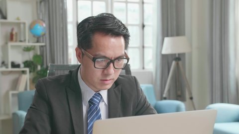 Asian Businessman With Glasses Wearing Business Suit Typing On Computer While Working At Home. 
