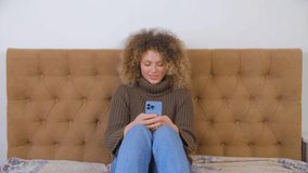 Happy white woman using mobile phone for communication. Beautiful curly female typing message online on social media app in blue smartphone. Cheerful Caucasian person use modern cellphone at home