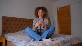 Happy young woman typing message on smartphone. Beautiful curly female using modern mobile phone for communication while sitting in bedroom at home during lockdown. Cheerful person messaging online