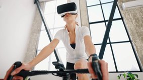 Young female in sports clothes and VR glasses is pedaling indoor cycle trainer. Studio flat, kitchen. Sunny day. Daily training at home. Modern technologies. Close up, slow motion, low angle shot