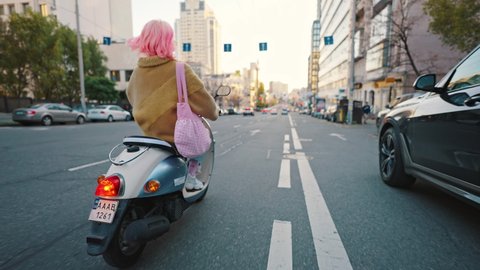 Freedom of movement. Follow shot of young trendy woman with pink hair riding on electric scooter along city street, back view 스톡 비디오