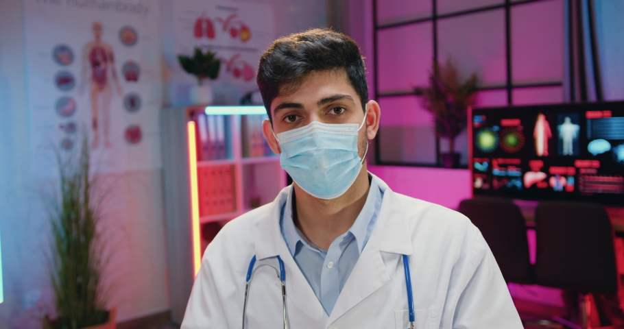 Close up portrait of likable confident skilled male chemist in medical uniform and facial mask which putting on his eyewear when looking into camera in dark lighted lab | Shutterstock HD Video #1086067538