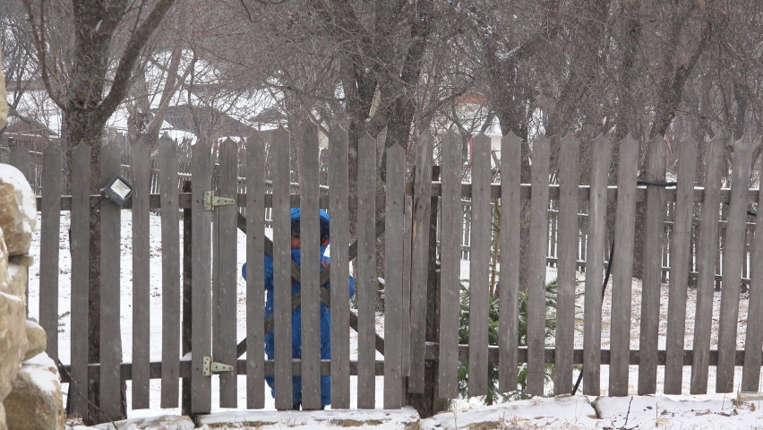 Little child comes through wooden rustic fence door into the backyard on snowy winter day