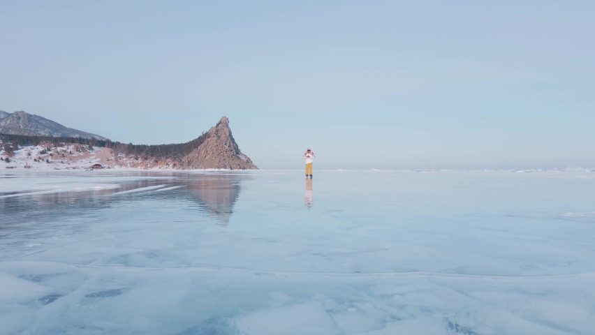 Aerial view of man skating on lake Baikal covered by ice. Male sportsman ejoying sport in cold weather with sky and clouds reflected in beautiful icy surface of lake.