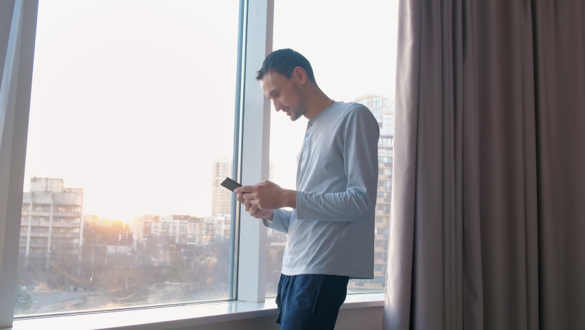 Young man is texting with his mistress at home by window. Wife approaches her husband and takes phone to see correspondence. Untrustworthy family relationships.