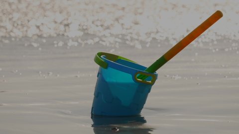 This serene telephoto video shows a blue child's sand pail on beach sand with slow motion waves crashing in the background.