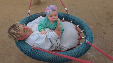 Woman with baby, lifestyle outdoors. Happy mother riding swing with cute little kid in urban park. Relaxation in free time. Red ropes, swing movement.