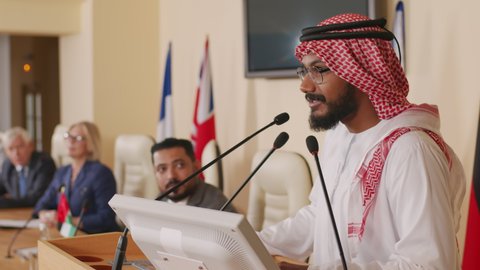 Waist up shot of male Arab politician in traditional clothes performing with speech on tribune at big conference hall with flashing cameras from press and other politicians listening to him