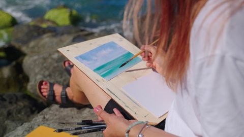 Woman artist painting marine scene with water colors sitting on the rock by the sea, slow motion