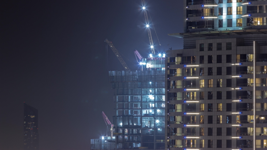 Tall buildings under construction and cranes in downtown night timelapse. Work progress at construction site of new towers behing skyscraper with glowing windows