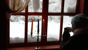 Winter house with a woman sitting on the terrace by a wooden window with snow on the glass drinking hot coffee with steam in warm clothes.Clip art concept calm secluded atmosphere with burning candle