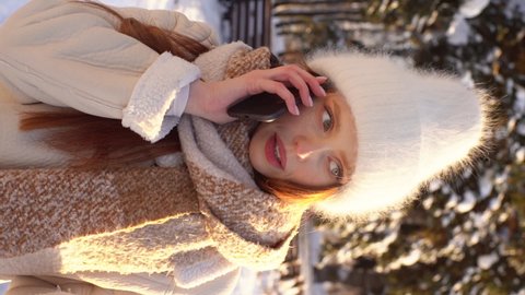 Vertical portrait of cheerful young woman in warm winter clothes talking on mobile phone outdoors in sunny winter day. Front view of smiling female using smartphone in snowy city park, slow motion.